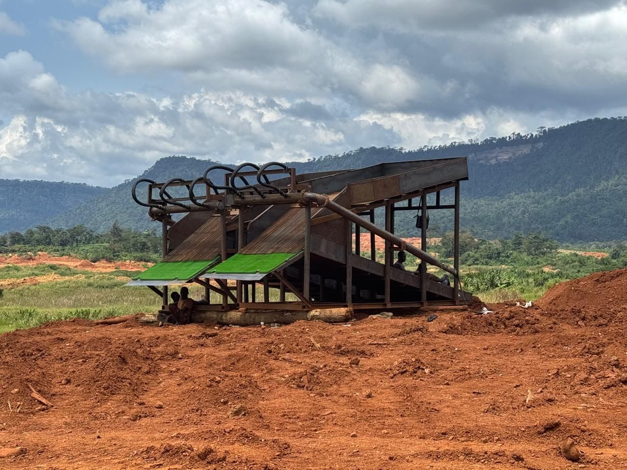 A gold processing plant at a sourcing site: steel sluice structure on red earth, with forested hills behind.